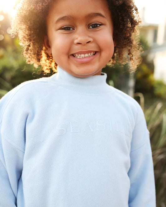 A smiling young child with curly hair wearing a light blue mock-neck sweatshirt with the 'SEASIDE' logo embossed on the chest, set against a warm, sunlit outdoor background.