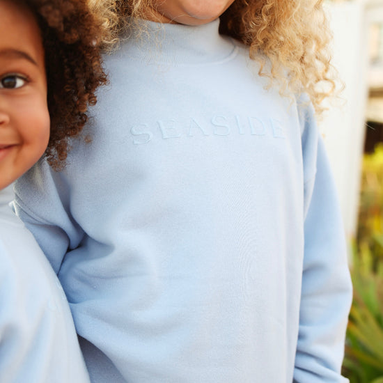 A close-up lifestyle shot of a child wearing a light blue mock neck sweatshirt with 'SEASIDE' embossed on the chest, with another child partially visible on the left.