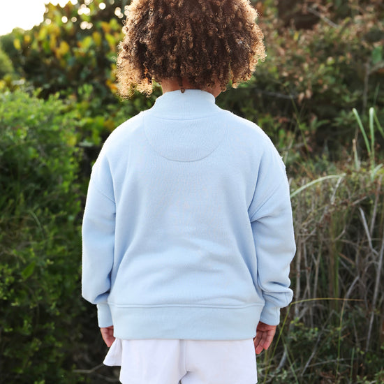 A child viewed from behind wearing a light blue sweatshirt and white shorts, standing outdoors in front of green foliage.