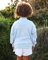 A child viewed from behind wearing a light blue sweatshirt and white shorts, standing outdoors in front of green foliage.