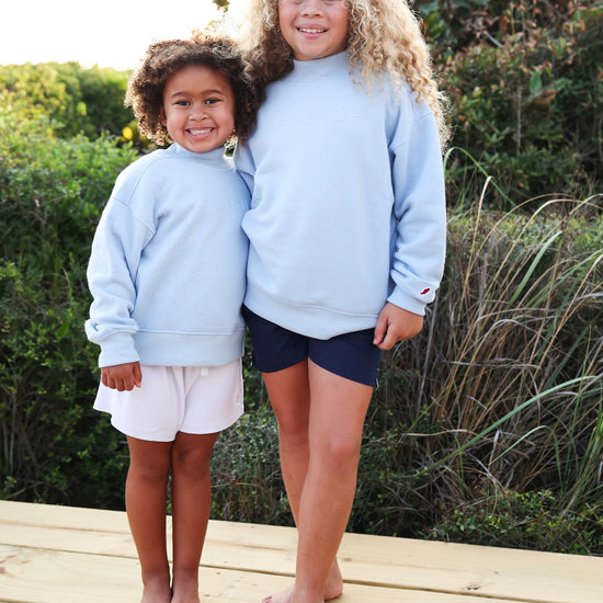 Two young girls standing barefoot on a wooden boardwalk outdoors, wearing matching light blue Seaside sweatshirts and different shorts (one white, one navy).
