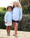 Two young girls standing barefoot on a wooden boardwalk outdoors, wearing matching light blue Seaside sweatshirts and different shorts (one white, one navy).