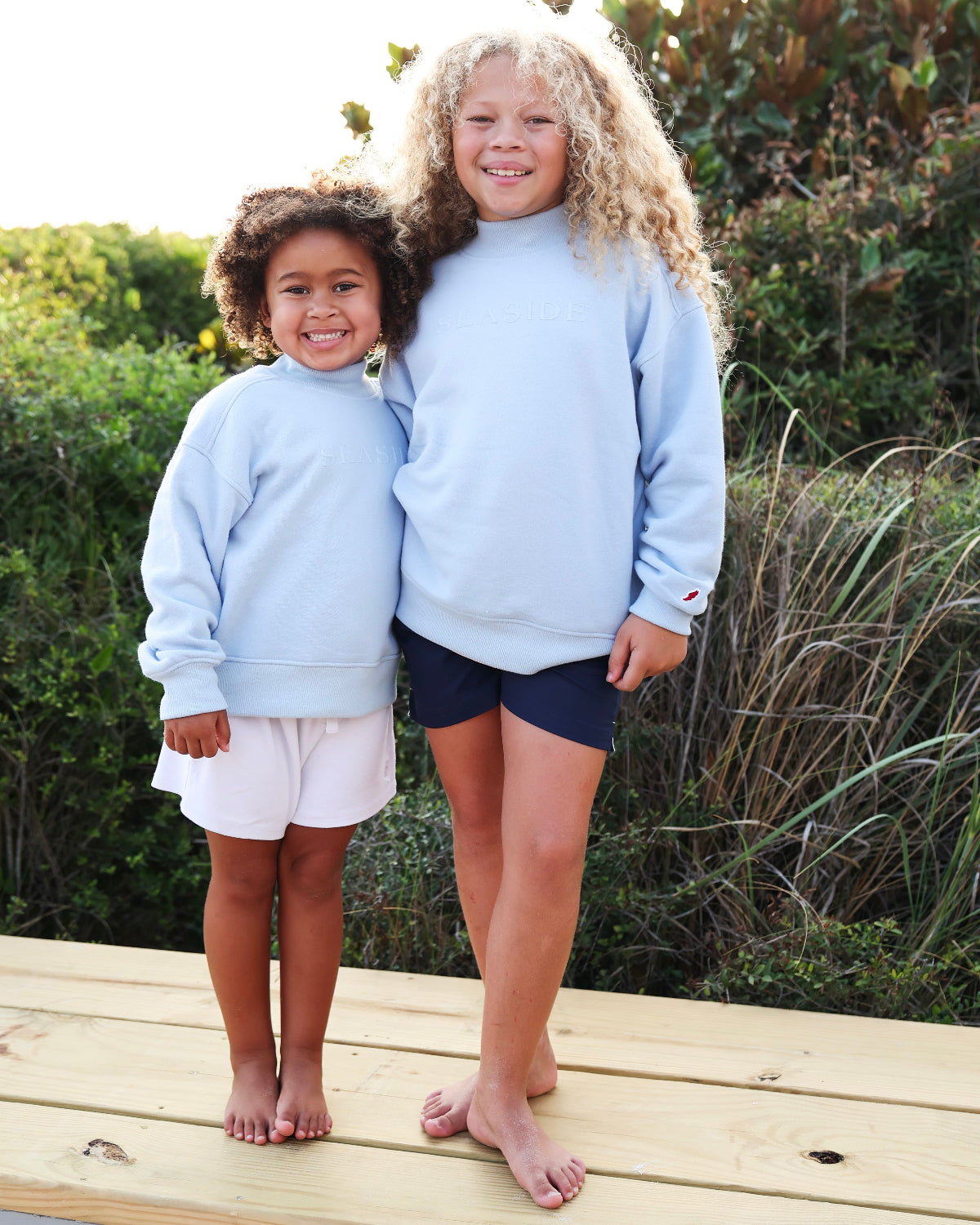 Two young girls standing barefoot on a wooden boardwalk outdoors, wearing matching light blue Seaside sweatshirts and different shorts (one white, one navy).