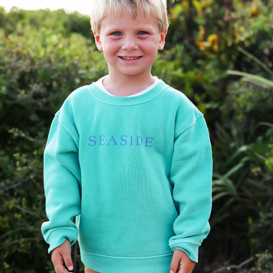 A young boy with blonde hair stands outdoors wearing a clover crewneck sweatshirt with the word 'SEASIDE' printed in blue across the chest, paired with tan pants.