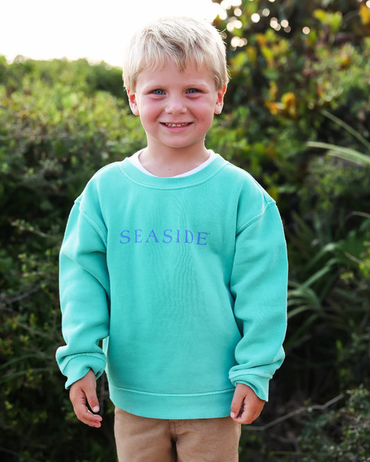 A young boy with blonde hair stands outdoors wearing a clover crewneck sweatshirt with the word 'SEASIDE' printed in blue across the chest, paired with tan pants.