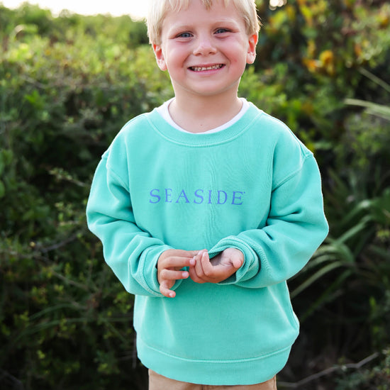 A young boy with blonde hair smiling outdoors, wearing a clover green crewneck sweatshirt with the word 'SEASIDE' printed in blue across the chest, paired with tan pants.