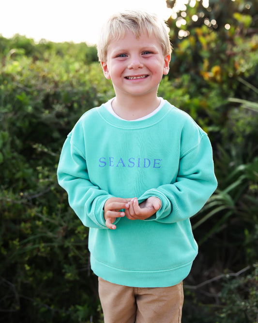 A young boy with blonde hair smiling outdoors, wearing a clover green crewneck sweatshirt with the word 'SEASIDE' printed in blue across the chest, paired with tan pants.