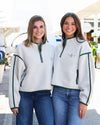 Two women standing side-by-side wearing matching off-white quarter-zip fleece pullovers with dark green trim and pocket details, paired with blue jeans.