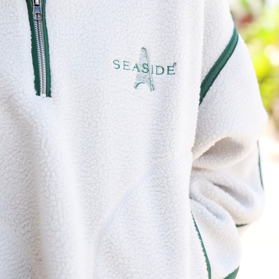 Close-up of a white fleece pullover jacket with green trim, a silver zipper, and 'SEASIDE' embroidered on the chest.