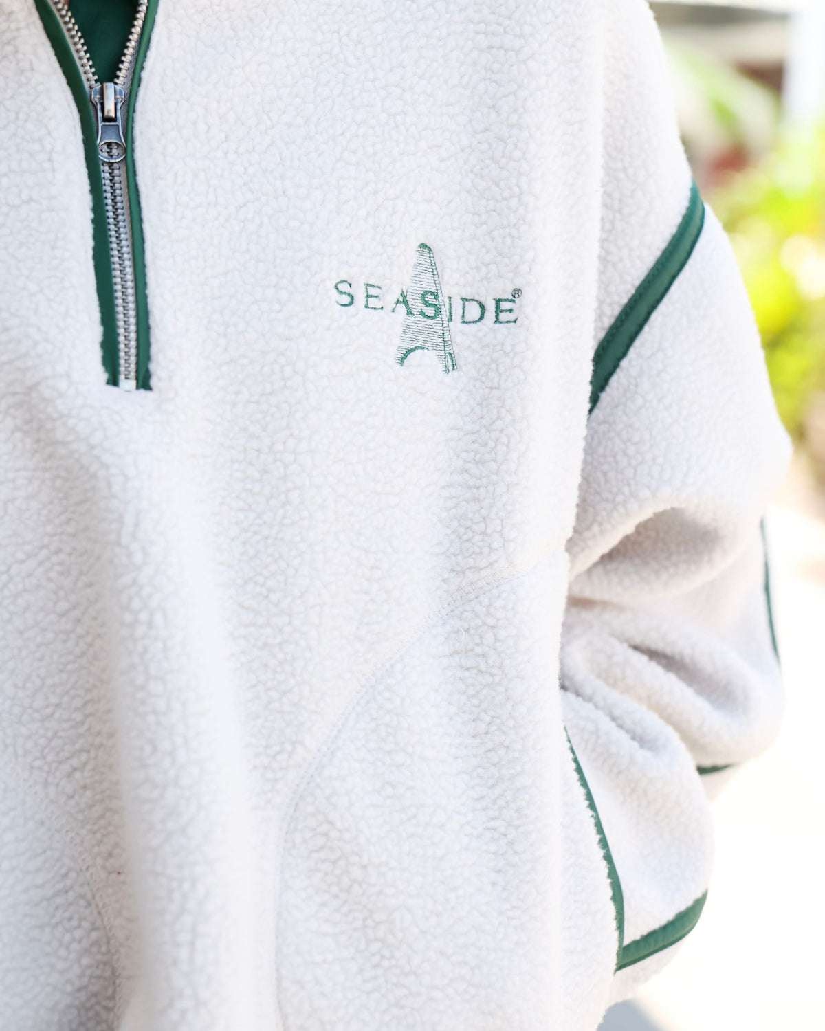 Close-up of a white fleece pullover jacket with green trim, a silver zipper, and 'SEASIDE' embroidered on the chest.