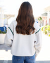 A woman viewed from behind wearing a cream-colored fleece pullover sweatshirt with green stripe accents on the shoulders, paired with blue jeans.