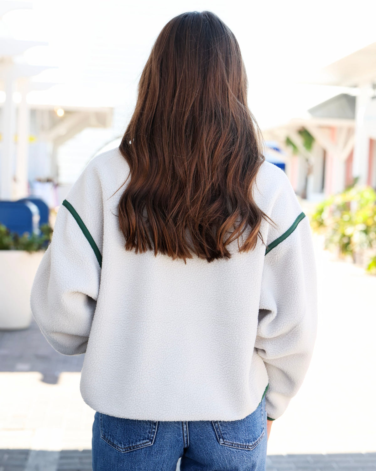 A woman viewed from behind wearing a cream-colored fleece pullover sweatshirt with green stripe accents on the shoulders, paired with blue jeans.