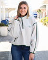 A smiling woman wearing a cream-colored fleece quarter-zip sweatshirt with dark green trim and a small embroidered logo on the left chest, paired with blue jeans, standing outdoors in a bright, sunny setting.