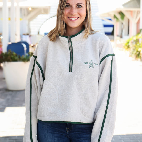 A smiling woman wearing a light gray fleece quarter-zip sweatshirt with dark green trim and 'SEA-MITE' printed on the chest, paired with dark blue jeans, standing outdoors on a sunny day.