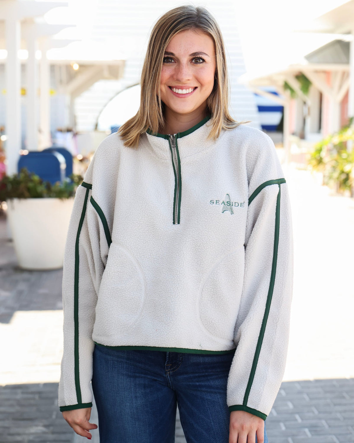 A smiling woman wearing a light gray fleece quarter-zip sweatshirt with dark green trim and 'SEA-MITE' printed on the chest, paired with dark blue jeans, standing outdoors on a sunny day.