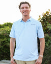 A smiling man wearing a light blue short-sleeve polo shirt with a subtle Coleman pavilion pattern and 'SEASIDE' printed on the chest, standing outdoors in front of green foliage.