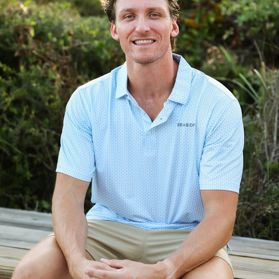 A smiling man sitting outdoors on a wooden bench wearing a light blue Coleman pavilion patterned short-sleeve polo shirt with a small visible SEASIDE logo on the chest, paired with khaki shorts, against a blurred green foliage background.