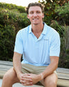 A smiling man sitting outdoors on a wooden bench wearing a light blue Coleman pavilion patterned short-sleeve polo shirt with a small visible SEASIDE logo on the chest, paired with khaki shorts, against a blurred green foliage background.