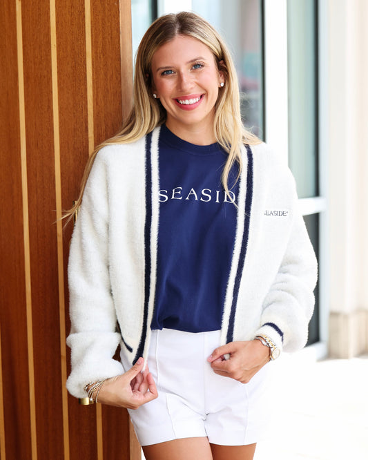 A smiling blonde woman wearing a fuzzy off-white cardigan with navy blue trim over a navy blue crewneck sweatshirt that says 'SEASIDE' and white shorts, standing in front of a wooden slatted wall.