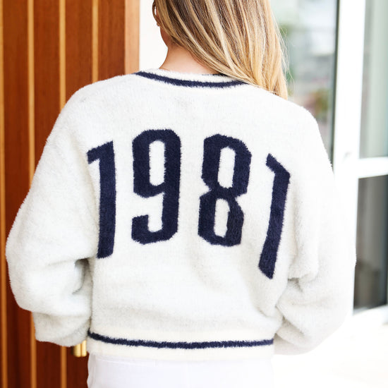 A woman viewed from behind wearing a fuzzy off-white sweater with '1981' in dark navy knit lettering across the back, paired with white pants.