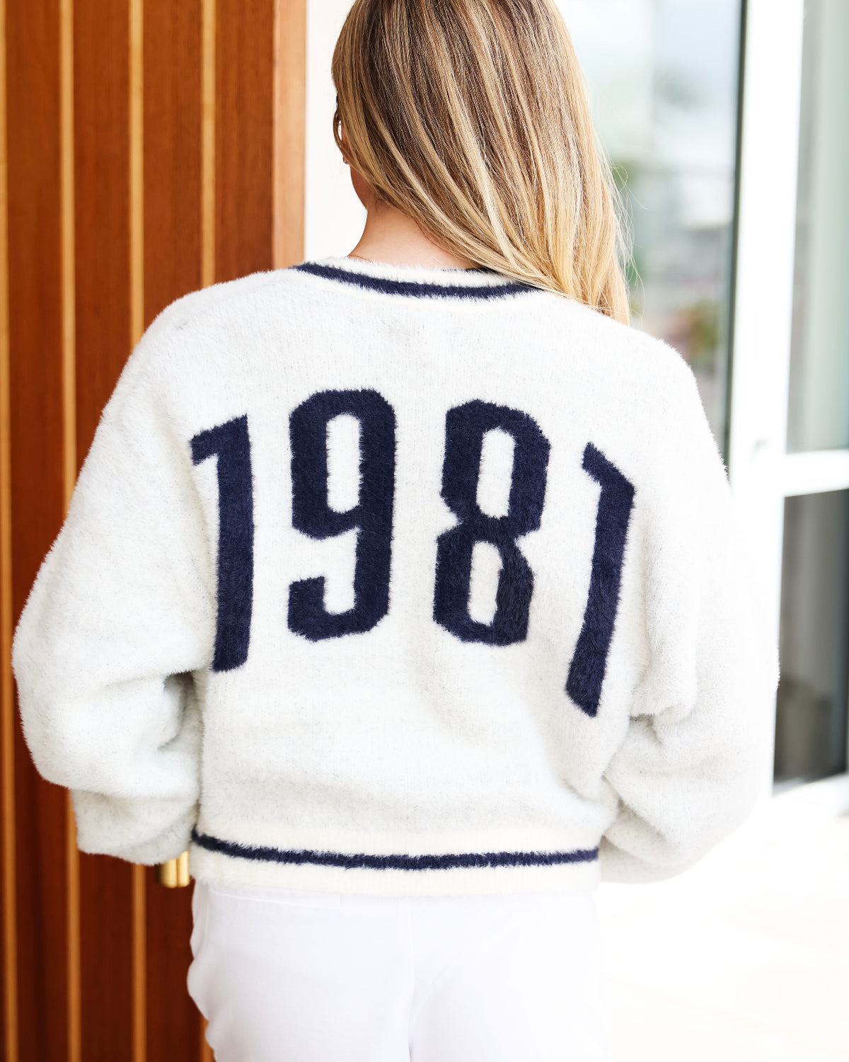 A woman viewed from behind wearing a fuzzy off-white sweater with '1981' in dark navy knit lettering across the back, paired with white pants.
