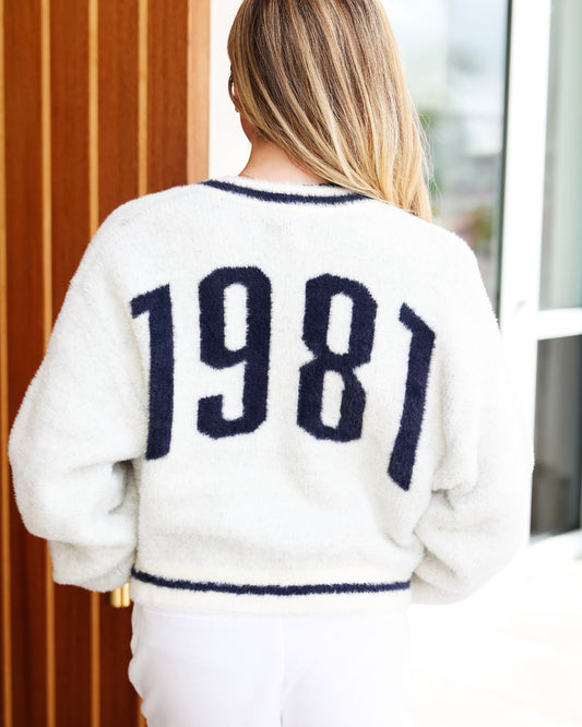 A woman viewed from behind wearing a fuzzy off-white sweater with '1981' in dark navy knit lettering across the back, paired with white pants.