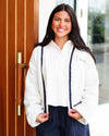 A smiling woman with long dark hair stands in front of a wooden door wearing a white fuzzy cardigan with dark navy trim and matching dark pants.