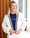A smiling blonde woman wearing a white fuzzy knit cardigan with navy blue trim over a navy blue crewneck sweatshirt with 'SEASIDE' printed in white, paired with white pants.