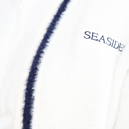 Close-up of a white textured cardigan with a navy blue fuzzy trim and 'SEASIDE' embroidered on the chest.