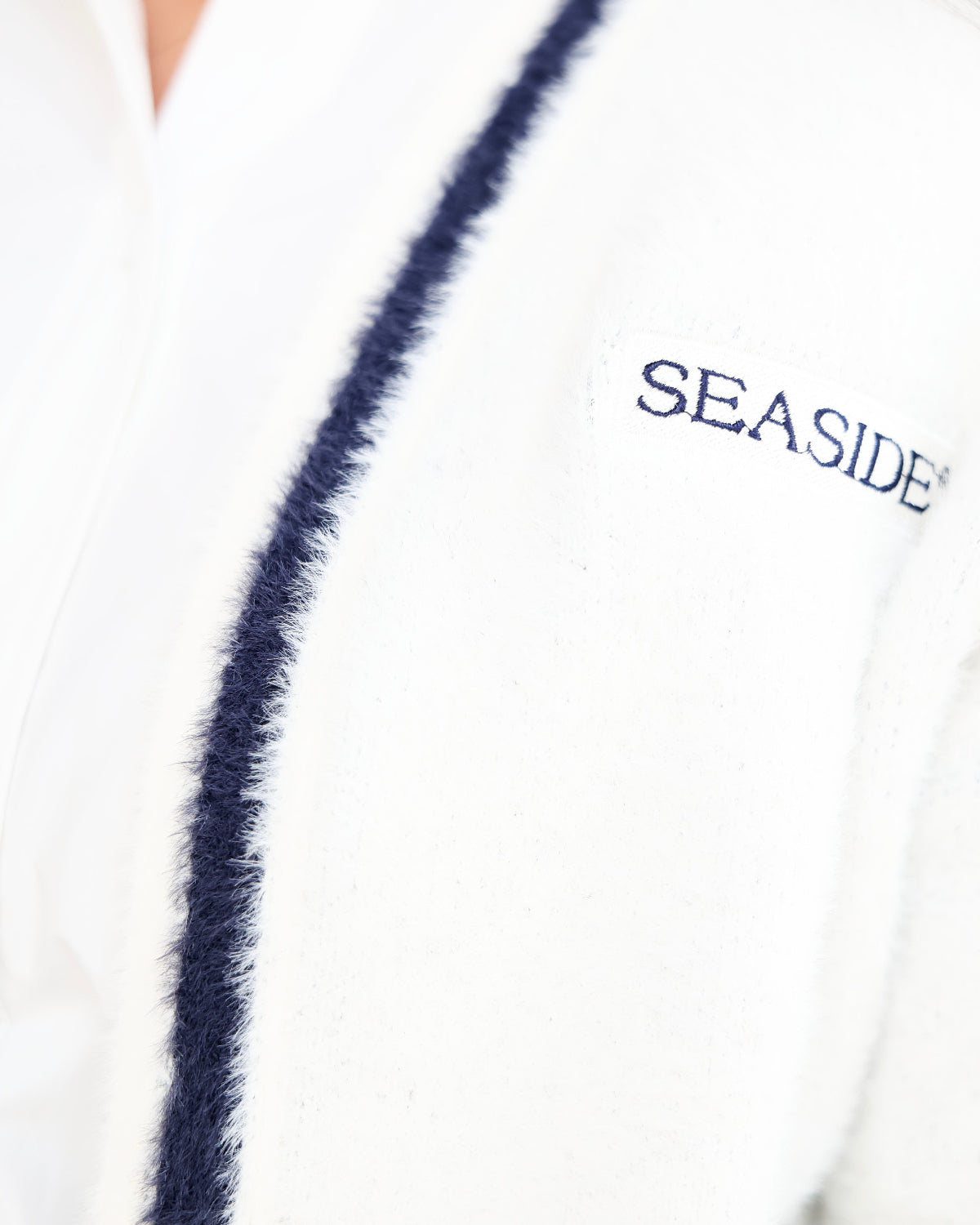 Close-up of a white textured cardigan with a navy blue fuzzy trim and 'SEASIDE' embroidered on the chest.