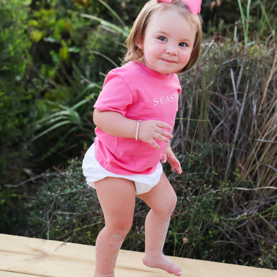 A toddler girl wearing a bright pink short-sleeve t-shirt with 'SEASIDE' printed on the chest, a matching pink bow in her hair, and a white diaper cover, standing barefoot on a wooden boardwalk outdoors.