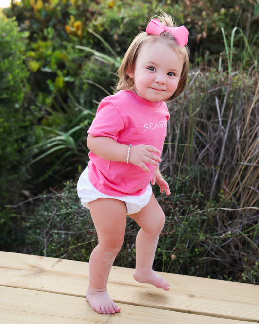 A toddler girl wearing a bright pink short-sleeve t-shirt with 'SEASIDE' printed on the chest, a matching pink bow in her hair, and a white diaper cover, standing barefoot on a wooden boardwalk outdoors.