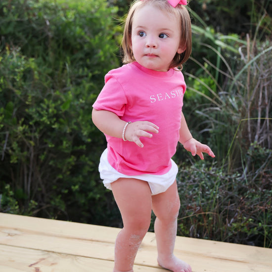 A toddler girl standing barefoot on a wooden deck wearing a bright pink short-sleeve t-shirt with 'SEASIDE' printed in white, white diaper cover, and a pink bow in her hair, with green foliage in the background.
