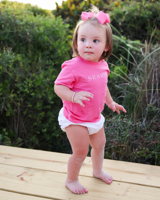 A toddler girl standing barefoot on a wooden deck wearing a bright pink short-sleeve t-shirt with 'SEASIDE' printed in white, white diaper cover, and a pink bow in her hair, with green foliage in the background.
