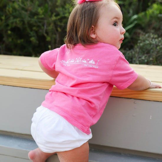 A toddler girl wearing a bright pink short-sleeve t-shirt and white shorts, climbing a wooden step outdoors. She has a matching pink bow in her hair.