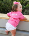 A toddler girl wearing a bright pink short-sleeve t-shirt and white shorts, climbing a wooden step outdoors. She has a matching pink bow in her hair.