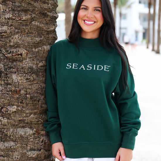 A woman smiling and leaning against a palm tree outdoors, wearing a dark green crewneck sweatshirt with 'SEASIDE' printed in white across the chest, paired with white shorts.