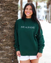 A woman smiling and leaning against a palm tree outdoors, wearing a dark green crewneck sweatshirt with 'SEASIDE' printed in white across the chest, paired with white shorts.