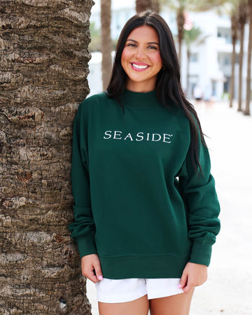 A woman smiling and leaning against a palm tree outdoors, wearing a dark green crewneck sweatshirt with 'SEASIDE' printed in white across the chest, paired with white shorts.