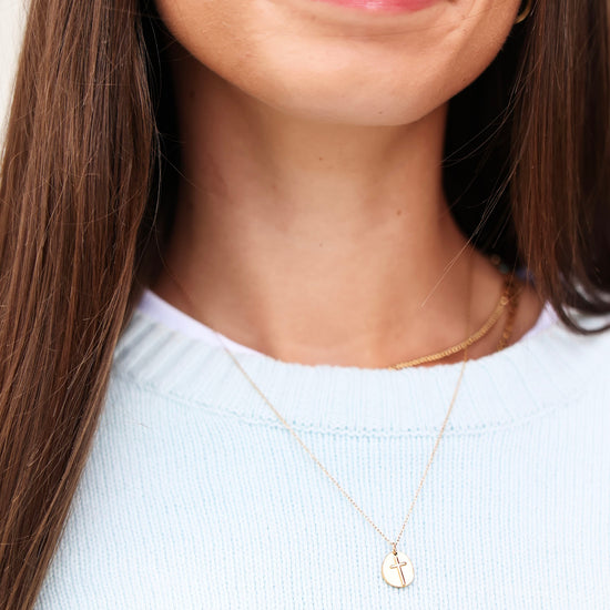 A close-up of a woman wearing a delicate gold necklace with a small oval cross pendant, paired with a light blue knit top.