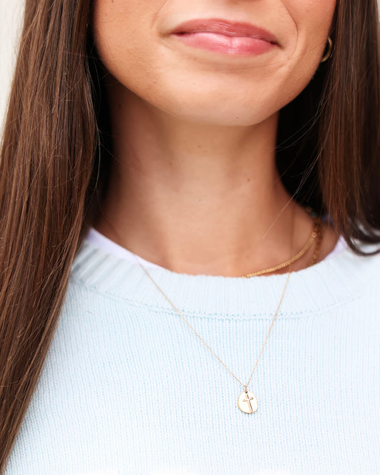 A close-up of a woman wearing a delicate gold necklace with a small oval cross pendant, paired with a light blue knit top.