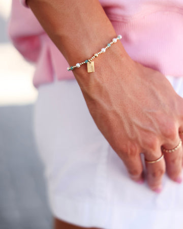 A delicate beaded bracelet with white pearls, turquoise beads, and a small gold seaside charm, worn on a woman's wrist.