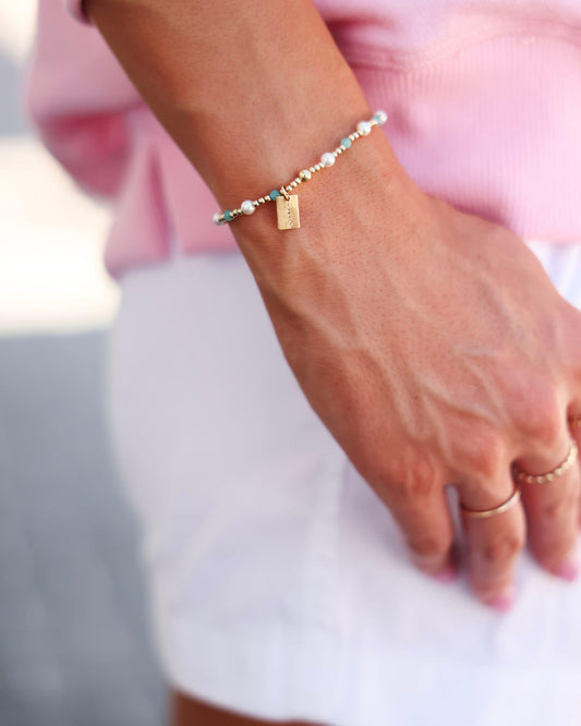 A delicate beaded bracelet with white pearls, turquoise beads, and a small gold seaside charm, worn on a woman's wrist.