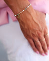A close-up of a person's hand wearing a delicate beaded bracelet with a small gold nameplate charm, alongside matching gold rings, against a soft pink and white background.