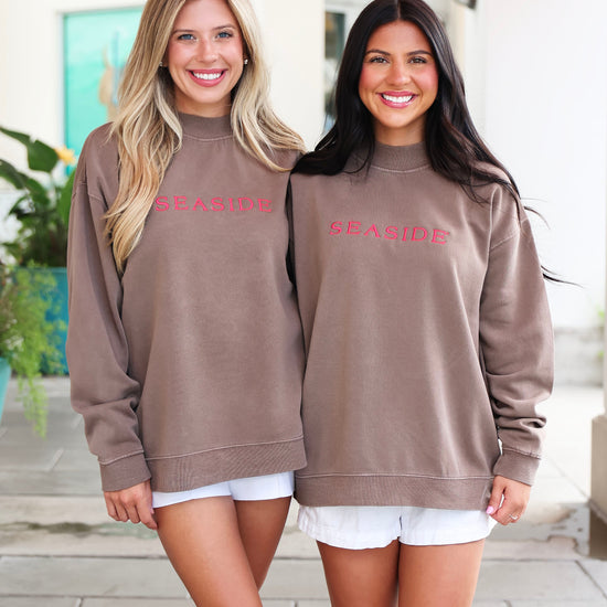 Two women standing side-by-side wearing matching taupe-brown crewneck sweatshirts with 'SEASIDE' printed in pink across the chest, paired with white shorts.