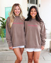 Two women standing side-by-side wearing matching taupe-brown crewneck sweatshirts with 'SEASIDE' printed in pink across the chest, paired with white shorts.