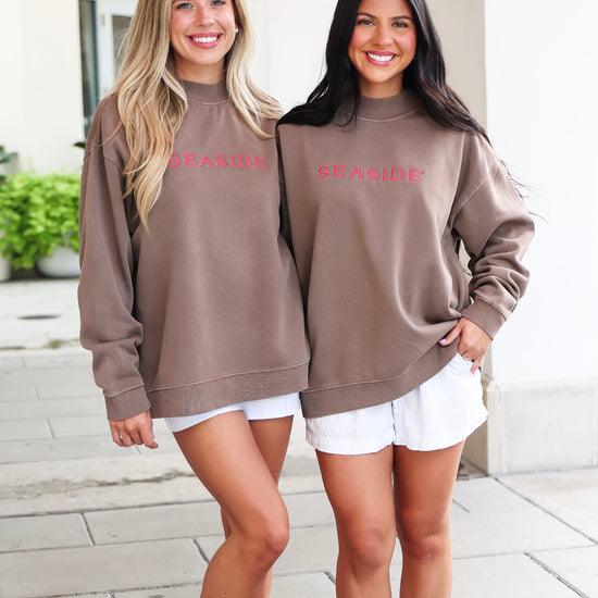 Two women standing outdoors on a paved walkway wearing matching taupe-brown sweatshirts with 'SEASIDE' printed in pink across the chest, paired with white shorts.
