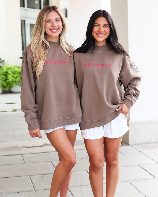 Two women standing outdoors on a paved walkway wearing matching taupe-brown sweatshirts with 'SEASIDE' printed in pink across the chest, paired with white shorts.