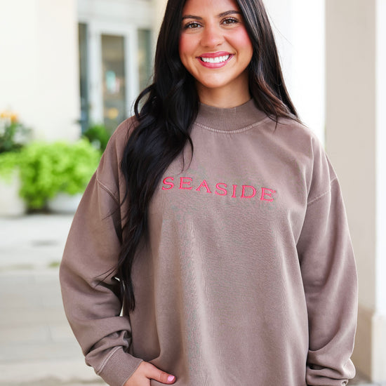 A smiling woman with long dark hair wearing an oversized taupe sweatshirt with 'SEASIDE' embroidered in pink across the chest, standing outdoors in front of a light-colored building.
