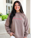 A smiling woman with long dark hair wearing an oversized taupe sweatshirt with 'SEASIDE' embroidered in pink across the chest, standing outdoors in front of a light-colored building.